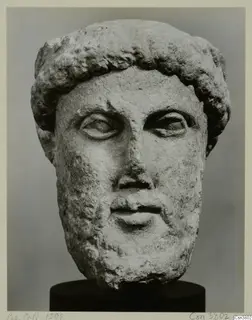 Carved stone head of a bearded man with short curly hair, shown on a dark cylindrical base.