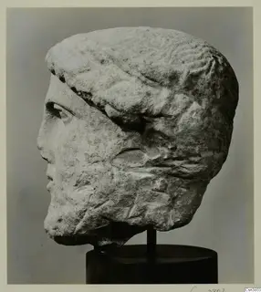Carved stone male head in profile, with a band across the hair, mounted on a black stand.