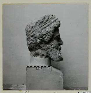 Carved stone head of a bearded man in profile on a square base, with a scale bar and number label.
