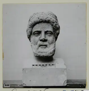 Carved stone head of a bearded man with curled hair and beard, set on a block base