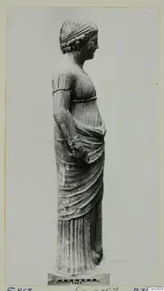 Standing stone statue of a draped female figure, shown in profile, with a measuring scale bar at its base.