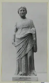 Stone statue of a standing female figure in a belted, draped dress, shown on a plinth with a scale bar.