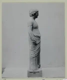 Standing female statue in stone, shown in profile facing right on a rectangular base.
