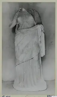 Standing draped statue in stone, head and neck missing, photographed against a grey backdrop