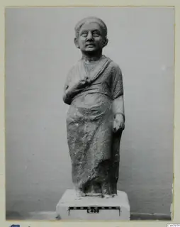 Standing stone statue of a draped figure on a white plinth, with a scale bar and number label in front.