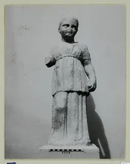 Carved stone statue of a standing figure in a belted, short-sleeved garment, on a base with a scale bar.