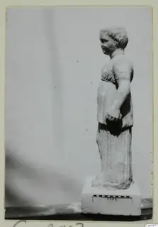 Pale stone statue of a standing figure in a belted tunic, shown in profile on a rectangular block base.