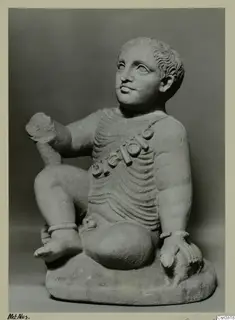 Stone statue of a seated child with a bead garland across the chest, photographed against a grey background.