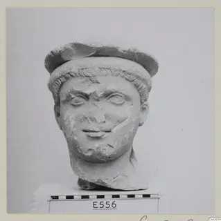 Carved stone head of a youth with a cap-like headdress, shown on a block against a plain background.