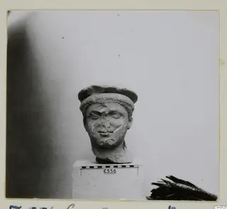Stone statue head with a cap-like headdress over short curled hair, set on a white plinth