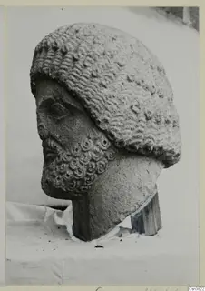 Carved stone male head with curly hair and beard, shown in side view against a pale backdrop.