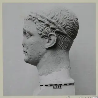 Stone head in profile facing left, with a band of leaves across curled hair, shown beside a scale bar and number label.