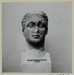 Carved stone head with curled hair, mounted on a square block, with a scale bar in front of the block.