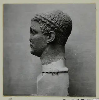 Carved stone head in profile, facing left, with curled hair and a band across the head, set on a plinth.