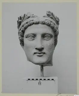 Carved stone head with curled hair and a leafy wreath, mounted on a rod above a plinth with a scale bar.