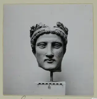 Carved stone head with curled locks and leaf-like ornaments above the hair, on a stand beside a scale bar.