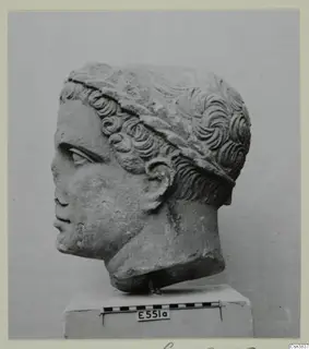 Carved stone head of a young figure in profile, with curly hair and a band across the head, on a plinth with a scale bar
