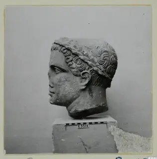 Carved stone head in left-facing profile with curled hair, set on a plinth with a scale bar.