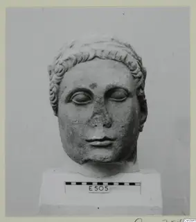 Carved stone head with curled hair and lowered eyes, standing on a plinth with a scale bar and number label.
