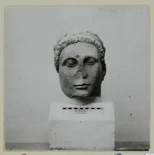 Carved stone head of a figure with curled hair, set on a rectangular block base with a scale bar