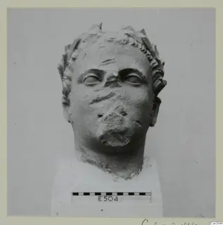 Stone head with a leafy wreath in the hair and a damaged nose and mouth, set on a white block base