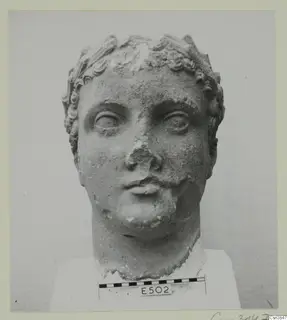 Stone head sculpture with curled hair and a raised band across the crown, shown front-on, nose broken away.