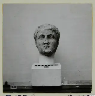 Stone head of a figure with curled hair, set on a square white plinth with a scale bar beside it.