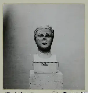 Stone head of a figure with curled hair and a broken nose, mounted on a square base in front of a plain backdrop.