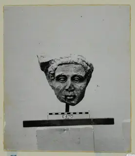 Carved stone head with curly hair band and wide eyes, shown on a wooden stand beside a scale bar and number label