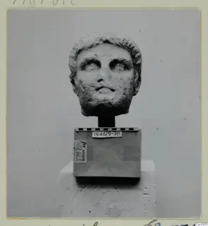 Stone sculpture of a head with short wavy hair, set on a box with a scale bar, photographed against a plain grey backdrop.