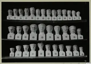 Three shelves of small pale sculpted heads on square white bases, each marked with a round number label.