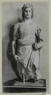 Stone statue of a draped standing figure with a bird perched on its right arm, set on a wooden base.