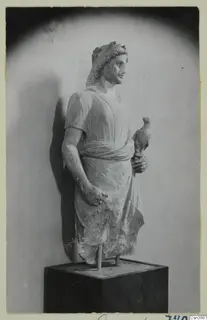 Stone statue of a standing male figure with a wreath in his hair, holding a small bird, on a dark wooden plinth