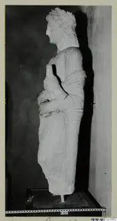 Stone statue of a standing figure in profile, with curled hair and a draped garment, on a dark wooden base.