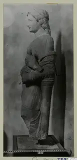 Stone statue of a female figure in left profile, with wavy hair and draped clothing, standing on a plinth.