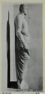 Standing draped stone statue of a figure seen in profile, facing right, on a plinth.