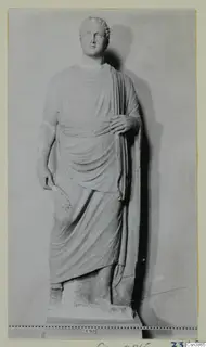 Standing stone statue of a draped figure with curled hair, photographed against a plain backdrop