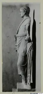 Standing stone statue of a draped figure, seen in side profile against a grey backdrop
