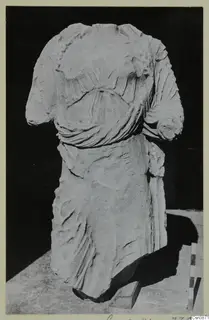 Headless marble statue torso of a draped figure, photographed against a dark background with a scale rod beside it.
