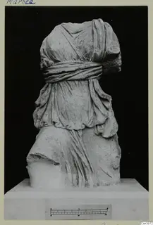 Headless draped statue in pale stone on a plinth, photographed against a black background, with a scale bar below