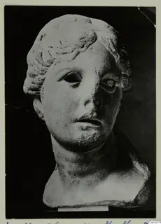 Marble head of a young woman with hair drawn back in waves and a bun, eyes hollow, nose broken.