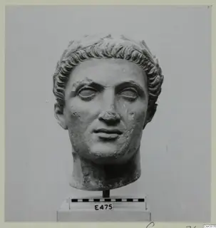 Stone head with short curled hair and a band across the top, mounted on a stand above a scale bar.