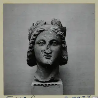 Carved stone head from Enkomi with curled hair and a leafy headdress, set on a block base
