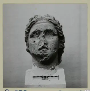 Stone head of a statue with wavy hair, shown on a plinth against a grey backdrop.