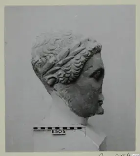 Stone head with curly hair and a wreath across the crown, shown in right profile against a grey backdrop