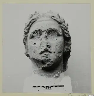 Carved stone head of a figure with wavy hair framing the face, eyes, nose and mouth worn.