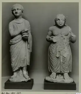 Two standing stone statues on dark bases; the left wears a draped garment and holds a round object, the right is bare-chested.