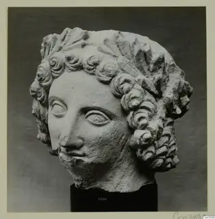 Marble head of a youth with curled hair, photographed on a black stand against a grey backdrop.