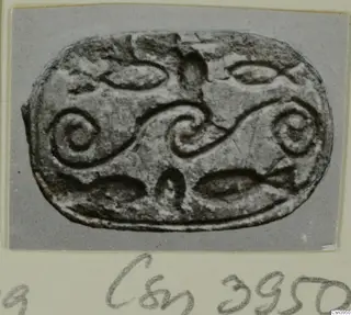 Oval seal or gem with raised spiral scrolls and curved leaf-like shapes, photographed face-on on a card mount.