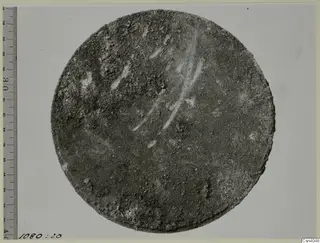 Round, corroded disc photographed beside a graduated measuring rod